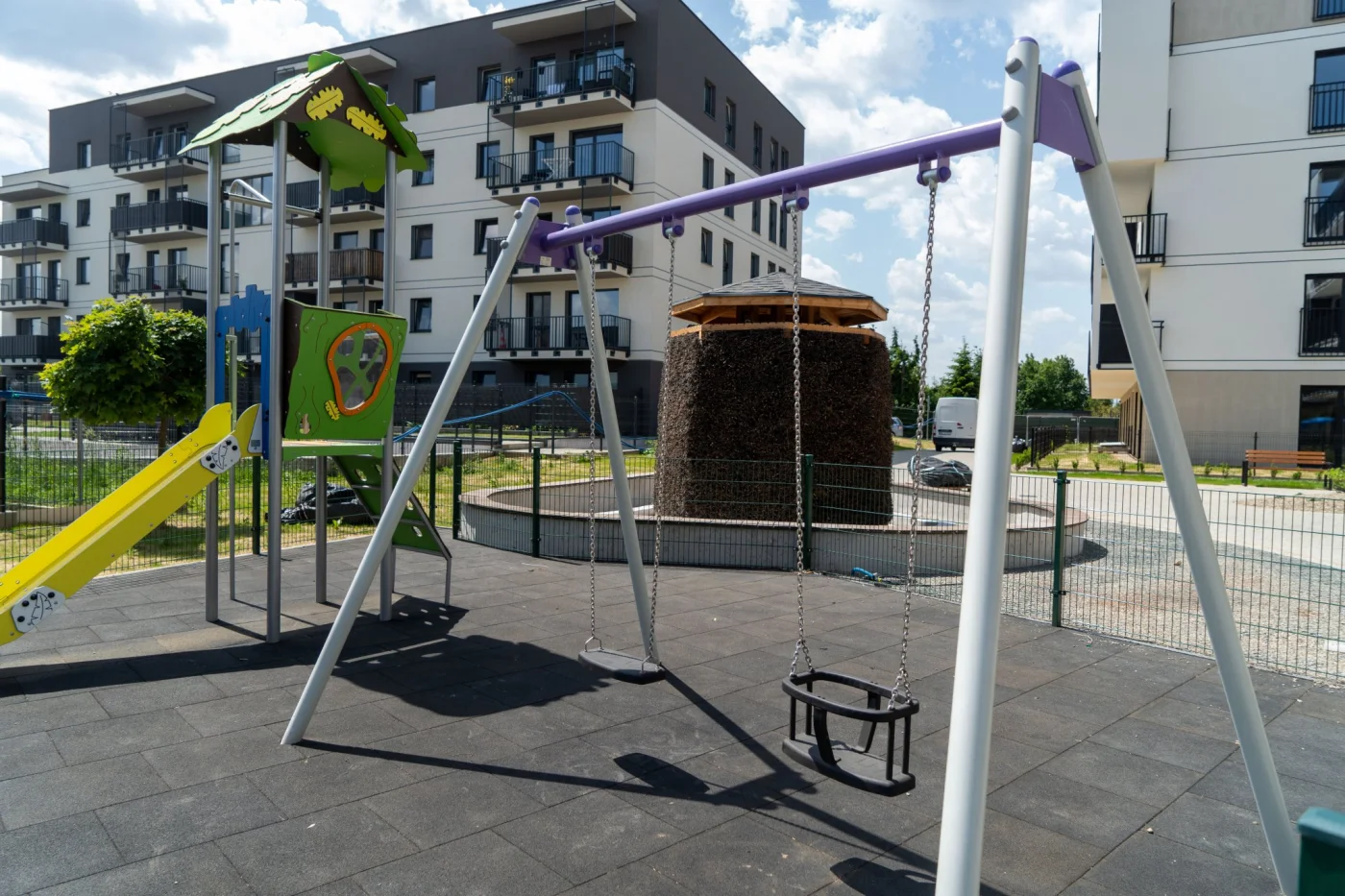 Plac zabaw z nowoczesnym sprzętem i elementami edukacyjnymi usytuowany przed budynkami mieszkalnymi w stylu współczesnej architektury.