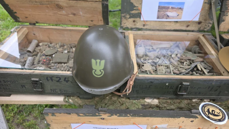 Zbliżenie na historyczny niemiecki stalowy hełm żołnierza Wehrmachtu z okresu II wojny światowej, umieszczony w drewnianej skrzyni obok amunicji i innych eksponatów wojskowych.