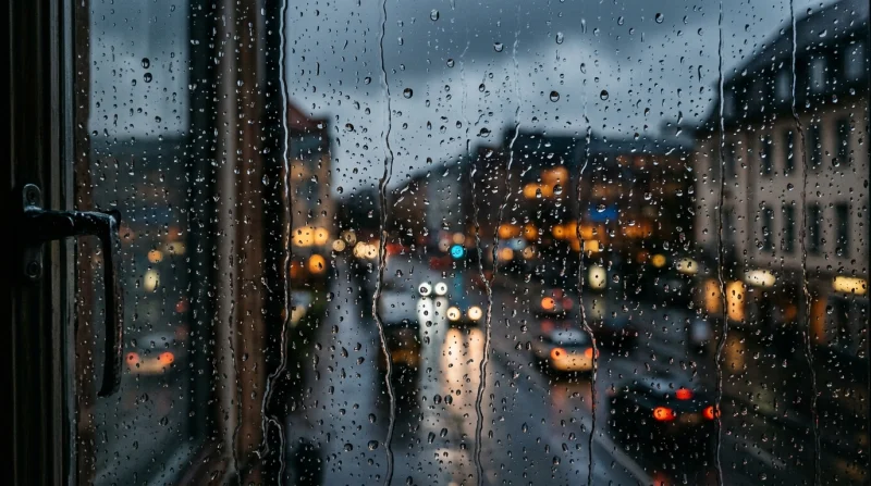 Widok ulicy za zaparowanym oknem pokrytym kroplami deszczu w świetle nocnych świateł miasta.