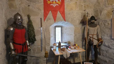 Scena historyczna przedstawiająca grupę rycerzy i postaci średniowiecznych w wnętrzu baszty lub zamku. Widoczne są elementy rekonstrukcji, takie jak zbroje, broń oraz dekoracje nawiązujące do epoki rycerskiej.