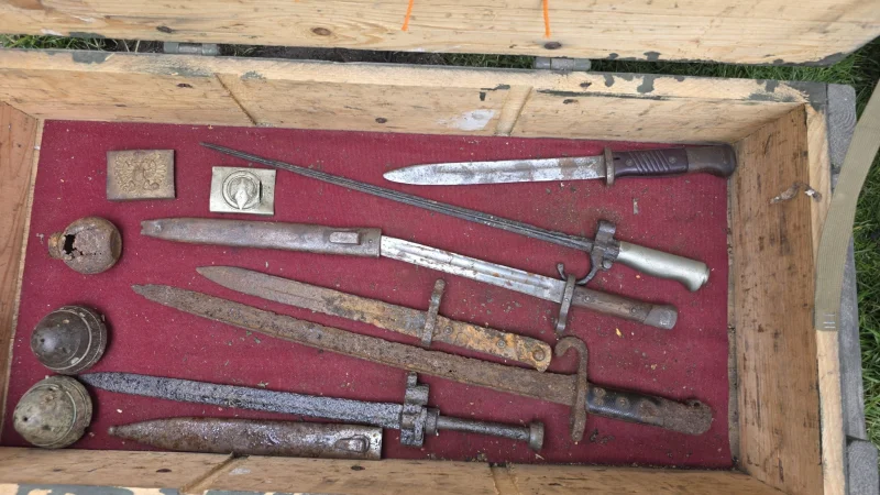 Zestaw starych narzędzi i przedmiotów metalowych, prawdopodobnie bagnety lub elementy historyczne, ułożony na czerwonym podkładzie w drewnianej skrzyni.